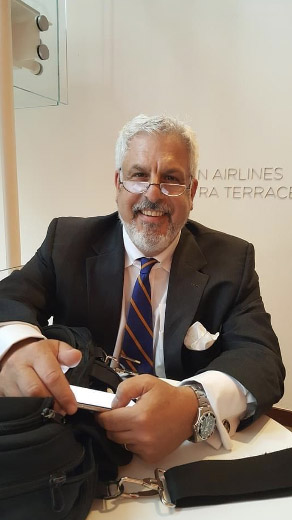 An image of Steven Cash posing in a black suit and tie while sitting at an airport.