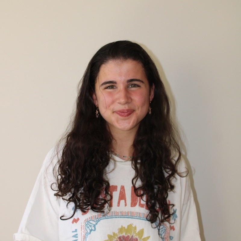 A person with long, curly dark hair stands in front of a light-colored wall, wearing a white t-shirt with a colorful floral and text design. They wear small dangling earrings and a thin necklace, smiling softly with a relaxed expression. Lighting is even and natural, with minimal shadows. The framing captures the upper half of the figure.