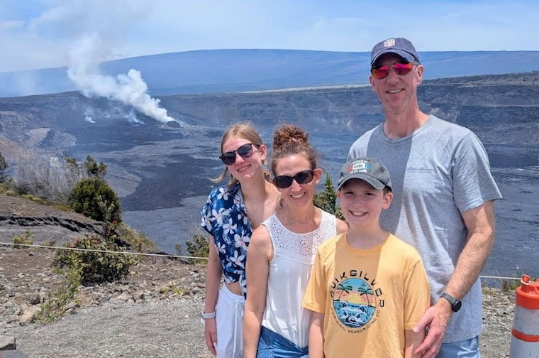  A group of four individuals poses for a photo at a volcanic crater with steam rising from it. They are standing on a paved lookout. The background shows a vast, rocky landscape under a hazy sky.