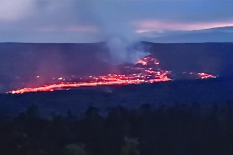 A glowing lava flow descends a hillside at dusk. The vibrant orange and red streaks of molten rock stand out against the dark landscape and smoky haze.