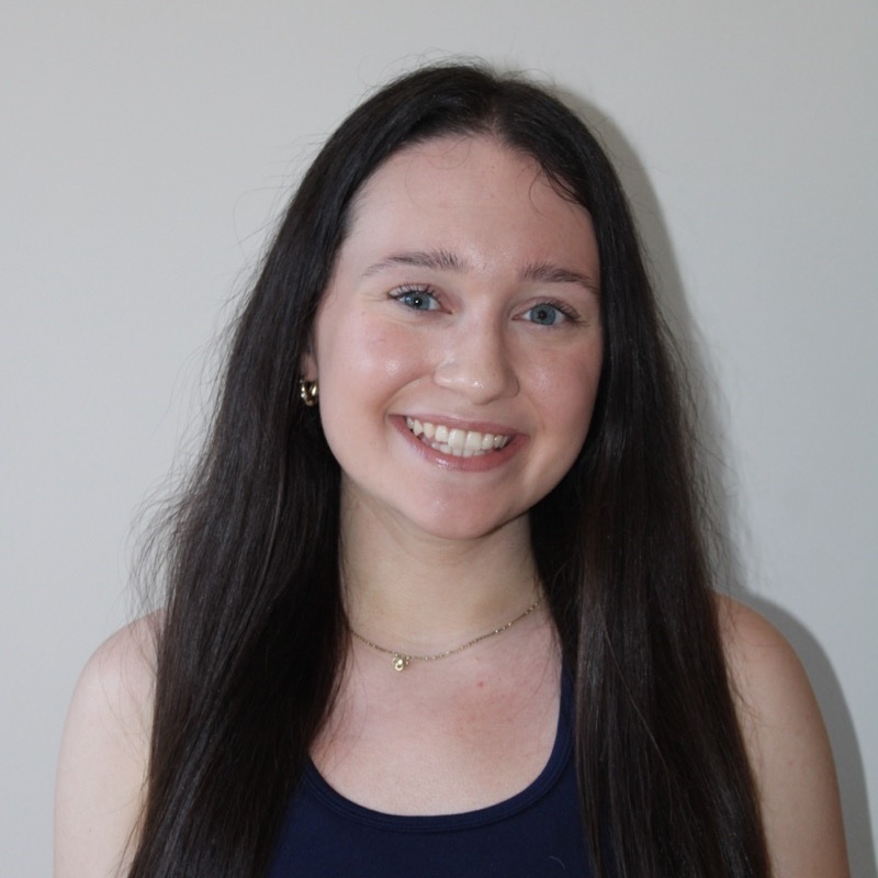 A person with straight, dark brown hair smiles directly at the camera. They wear a navy blue sleeveless top and a delicate chain necklace with a small charm. The lighting is soft and neutral, casting a cool tone. The background is light gray and uncluttered.