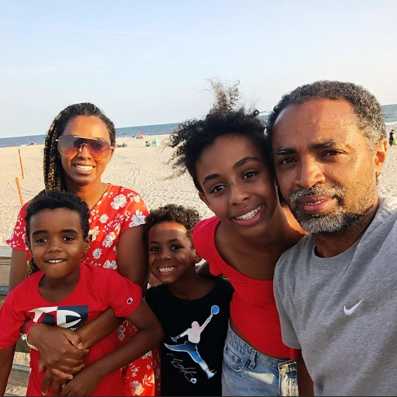 A group of five people poses for a selfie on a beach. They are all smiling and standing on a wooden walkway. The beach and ocean are visible in the background.
