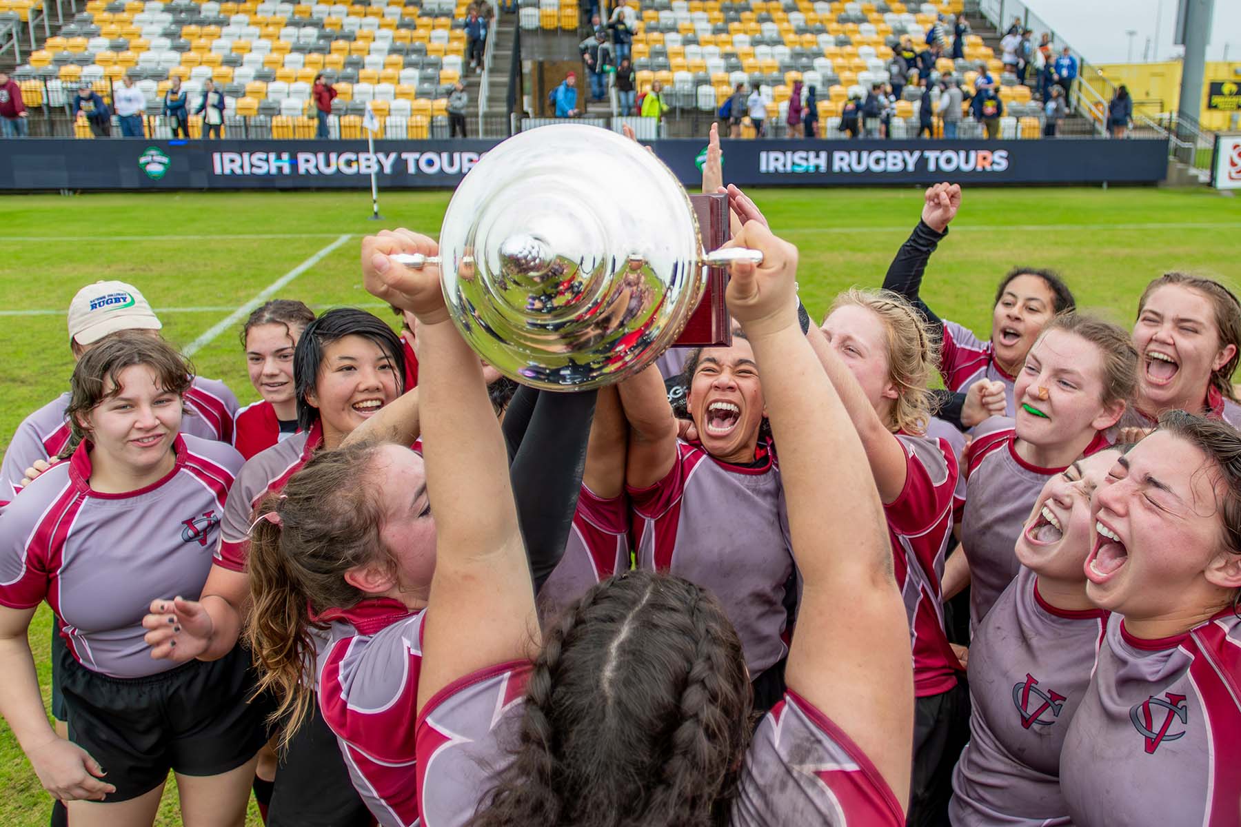Vassar College Women’s rugby team in maroon and gray jerseys celebrating on the field, holding up a large silver trophy together with excitement.