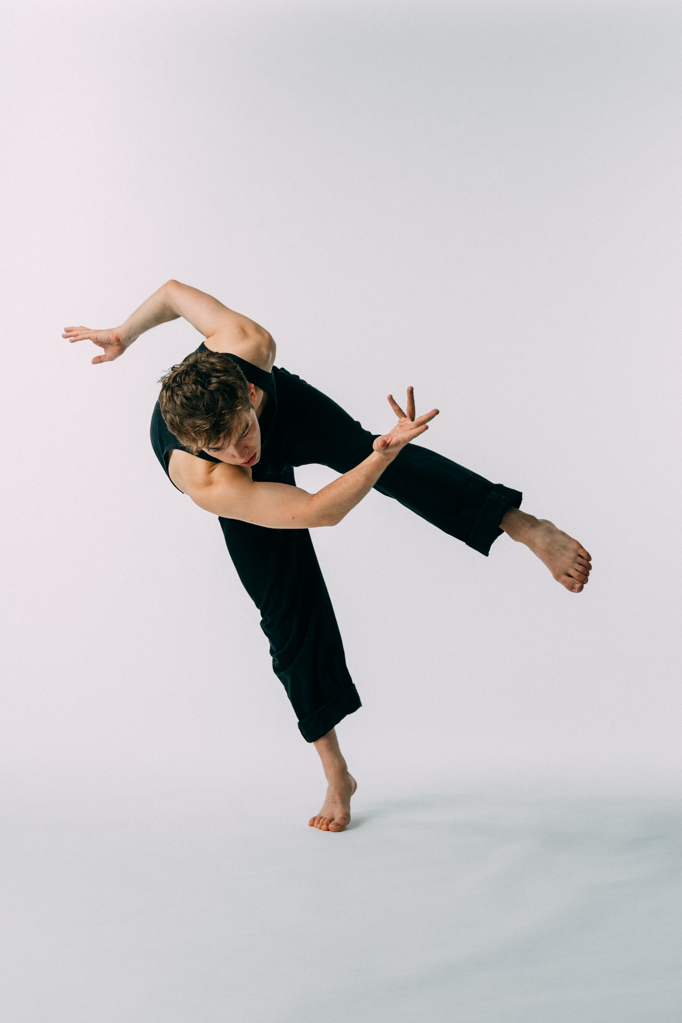 A person in a sleeveless black outfit performs a dynamic dance pose against a seamless white background. They balance on one foot with the other leg lifted and bent, while their torso leans forward and one arm extends outward in a curved motion. The fingers on one hand are splayed in a deliberate gesture, adding to the sense of motion. The lighting is even and soft, highlighting muscle definition and body alignment.