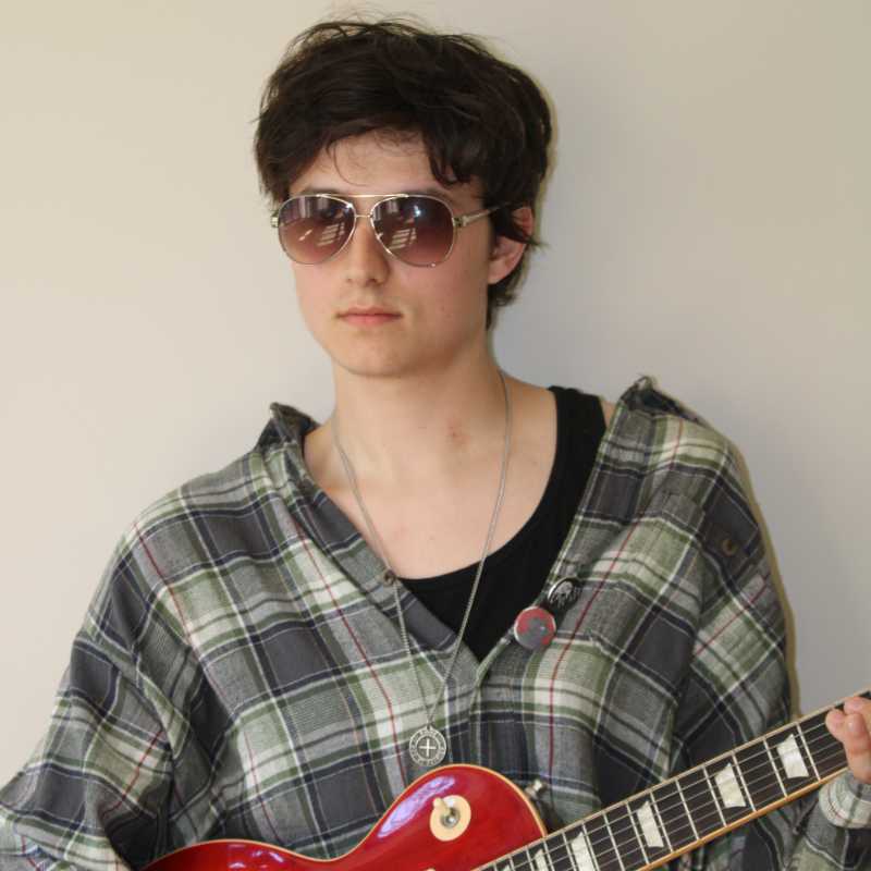 A person with tousled short hair wears large aviator sunglasses and a green plaid shirt layered over a black shirt. They hold a red electric guitar at chest height and wear a long pendant necklace. Their expression is neutral and relaxed. The background is a smooth, pale wall.