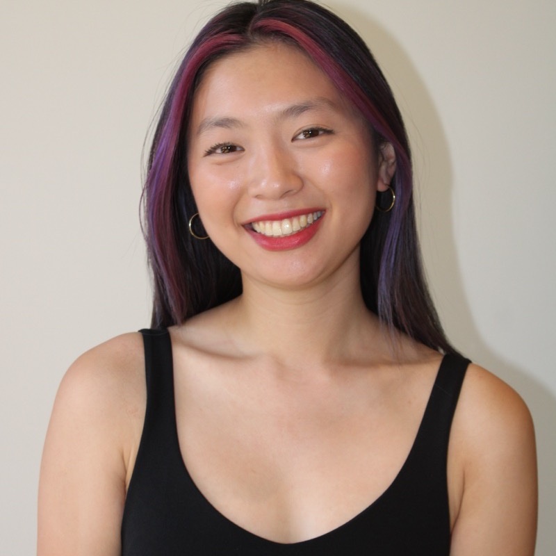 A person with long, straight hair dyed with pink and purple highlights smiles widely in front of a plain wall. They wear a black sleeveless top and gold hoop earrings. Their makeup is subtle, with bright red lipstick. Lighting is bright and even across the entire frame.