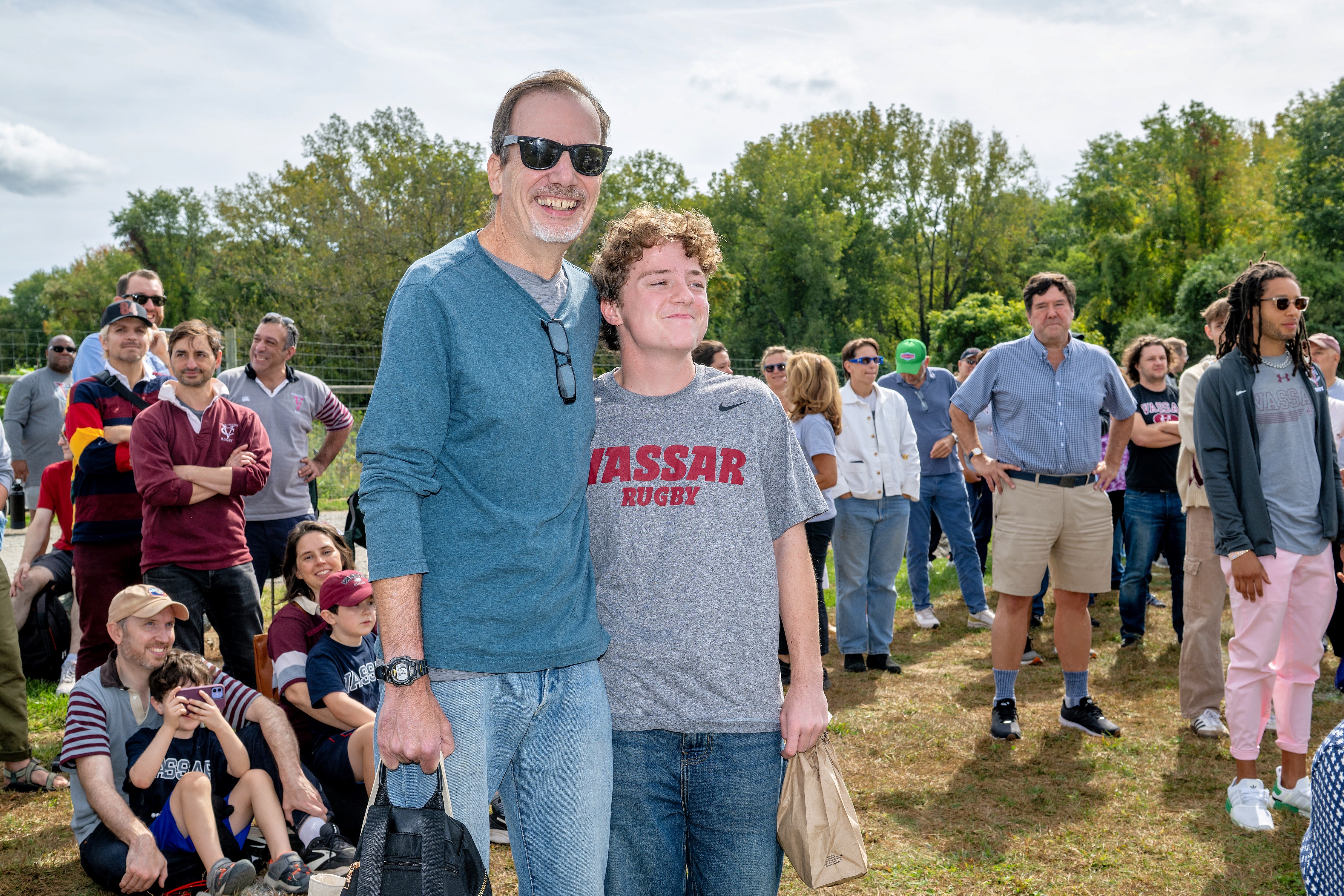A large group stands outdoors on a grassy field under a bright sky. In the foreground, two people pose closely together—one in a blue long-sleeve shirt and sunglasses, the other in a gray “Vassar Rugby” t-shirt holding a brown paper bag. Behind them, attendees stand and sit casually, many smiling or watching something out of frame. Trees form a green backdrop along the horizon.