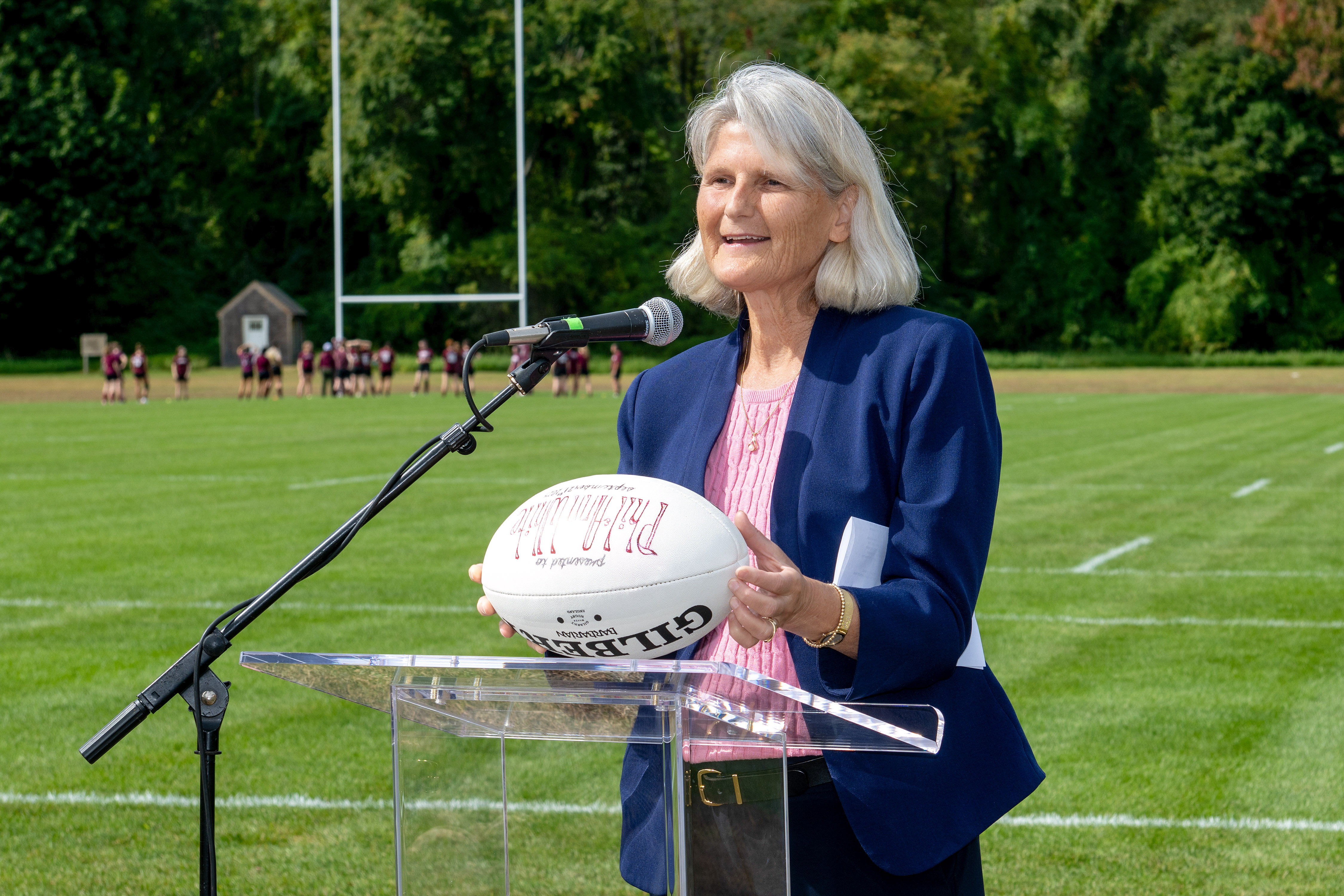 Elizabeth Bradley, a person with long gray hair, stands behind a clear podium with a microphone, holding a white rugby ball printed with red text. They wear a dark blue blazer and a pink knit shirt. Behind them, a rugby field stretches out with goalposts, short green grass, and a row of players standing near the edge. Trees line the background under a lightly overcast sky.