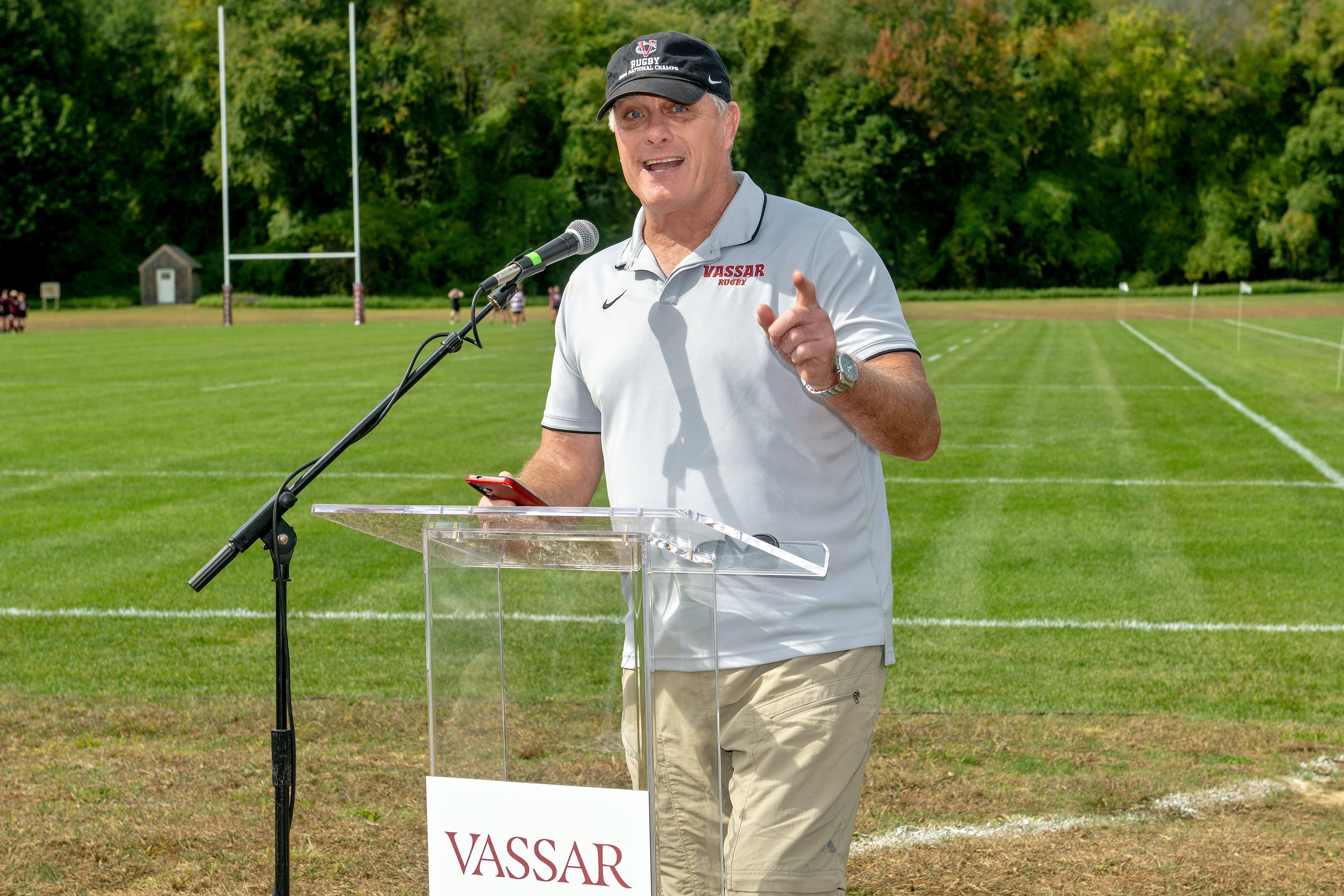 A person speaks at a clear podium with a microphone, pointing with one hand and holding a phone in the other. They wear a light gray “Vassar Rugby” polo shirt and a black cap with red and white embroidery. The rugby field extends behind them, bordered by goalposts and tall trees under a partly cloudy sky.