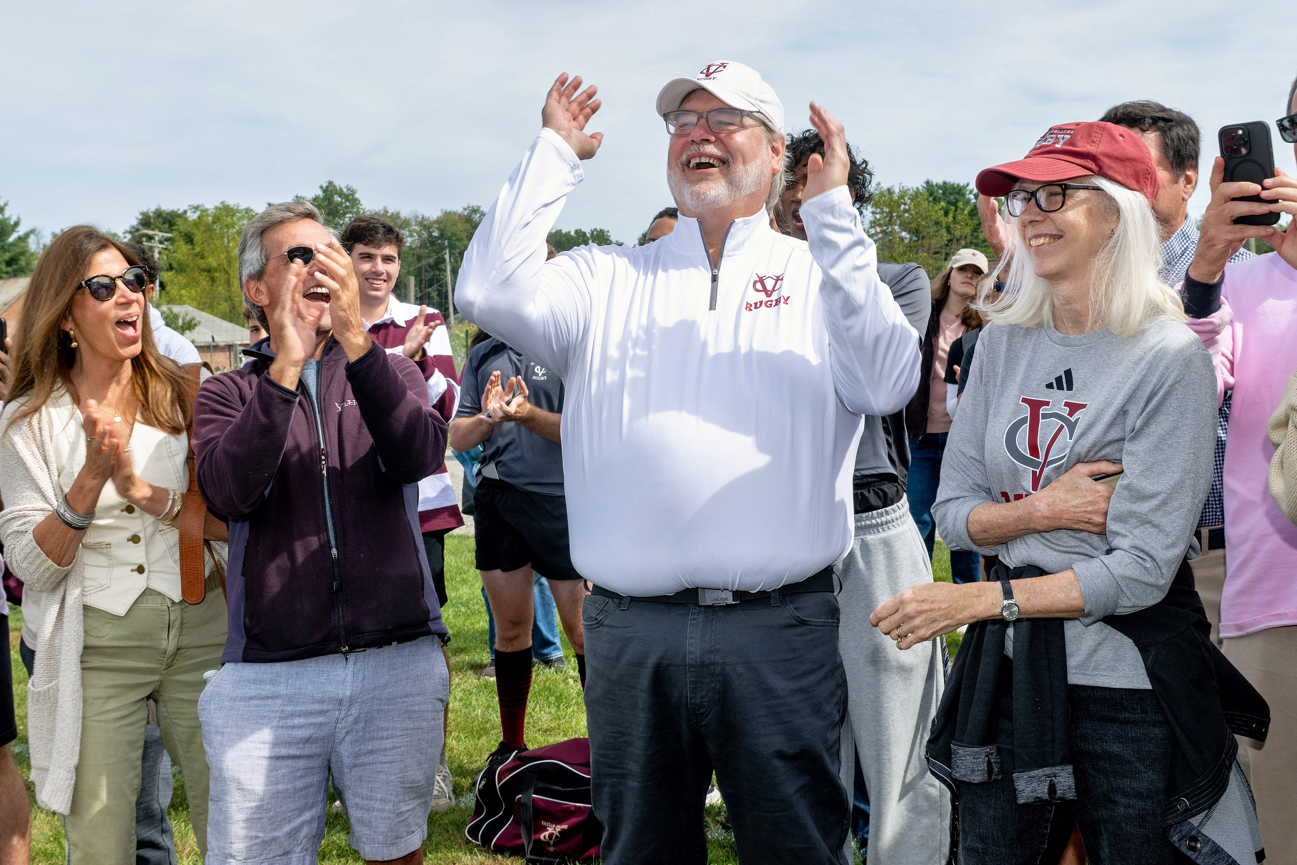 A crowd stands outdoors on grass, clapping and smiling. In the center, a person in a white quarter-zip pullover with a maroon “Rugby” logo raises both arms joyfully. People around them, some wearing sunglasses and Vassar-themed clothing, join in the celebration. The background includes trees and people partially visible in athletic uniforms.