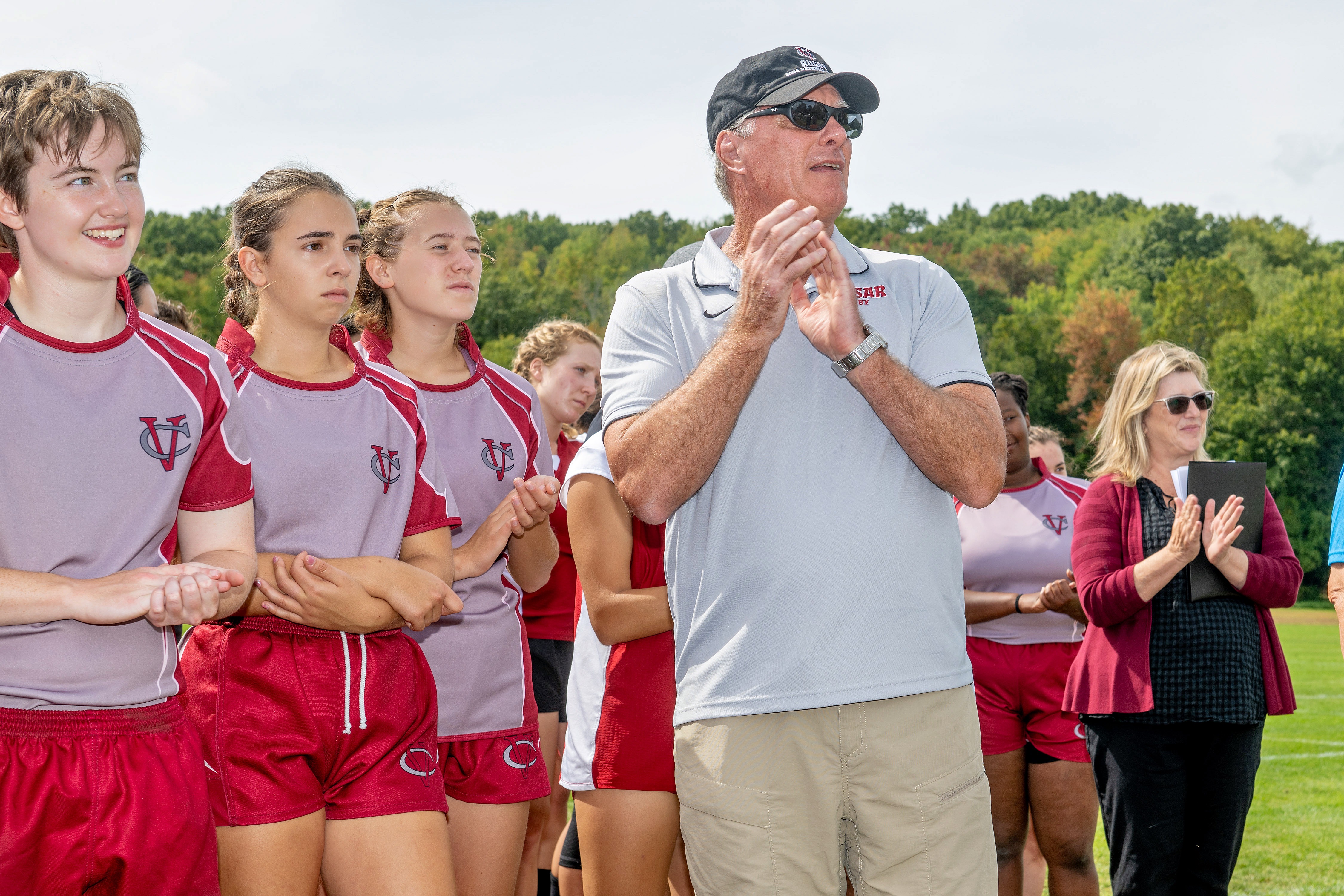 A group of people in red and gray athletic uniforms stands clapping near a rugby field. One person in front wears a “Vassar Rugby” polo and black cap, also clapping. Additional people, including someone in a maroon cardigan holding a tablet, are visible to the right. Trees and open grass form the background under a bright sky.