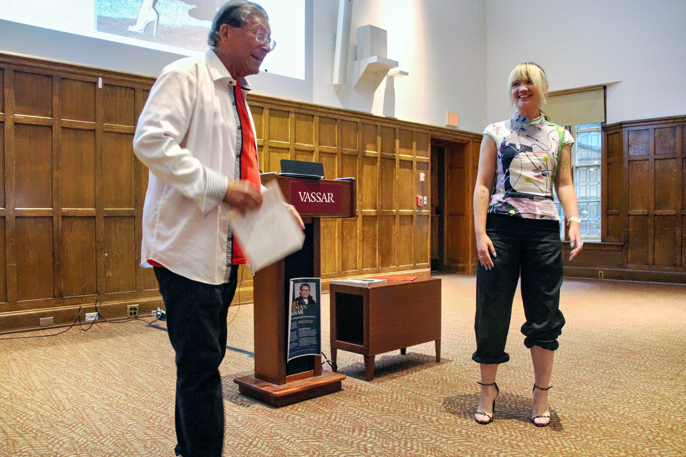 Two people standing before an audience on either side of a podium.