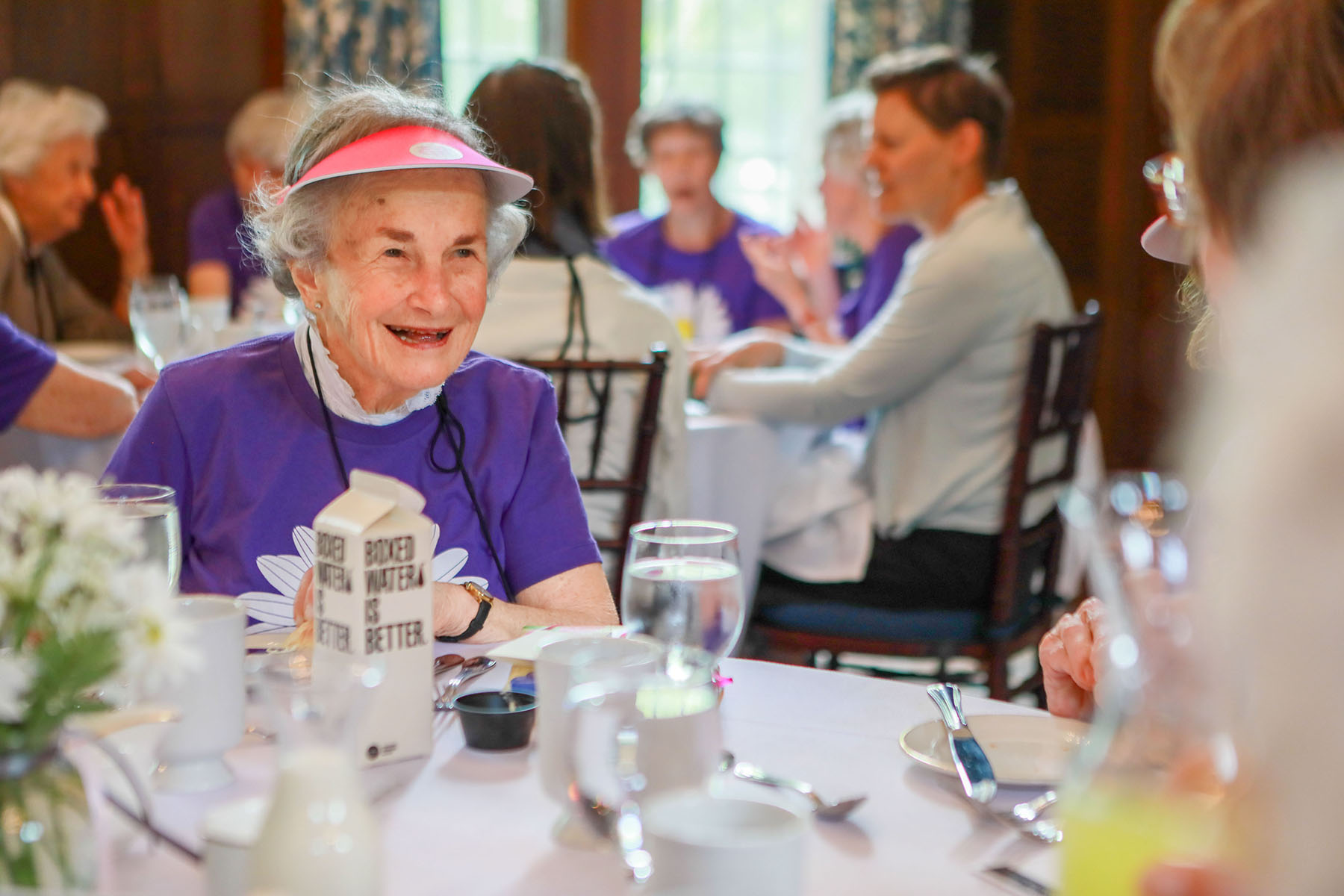 June Ross Marks ’49 with short white hair wearing a bright purple shirt and a pink visor, seated at a dining table.