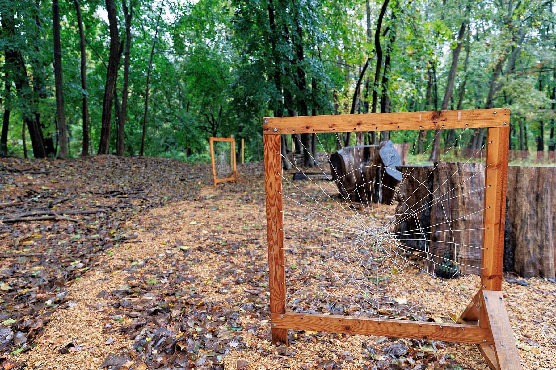 A nature-based learning exhibit in a wooded area, featuring two square wooden frames with string woven into a spiderweb-like pattern.