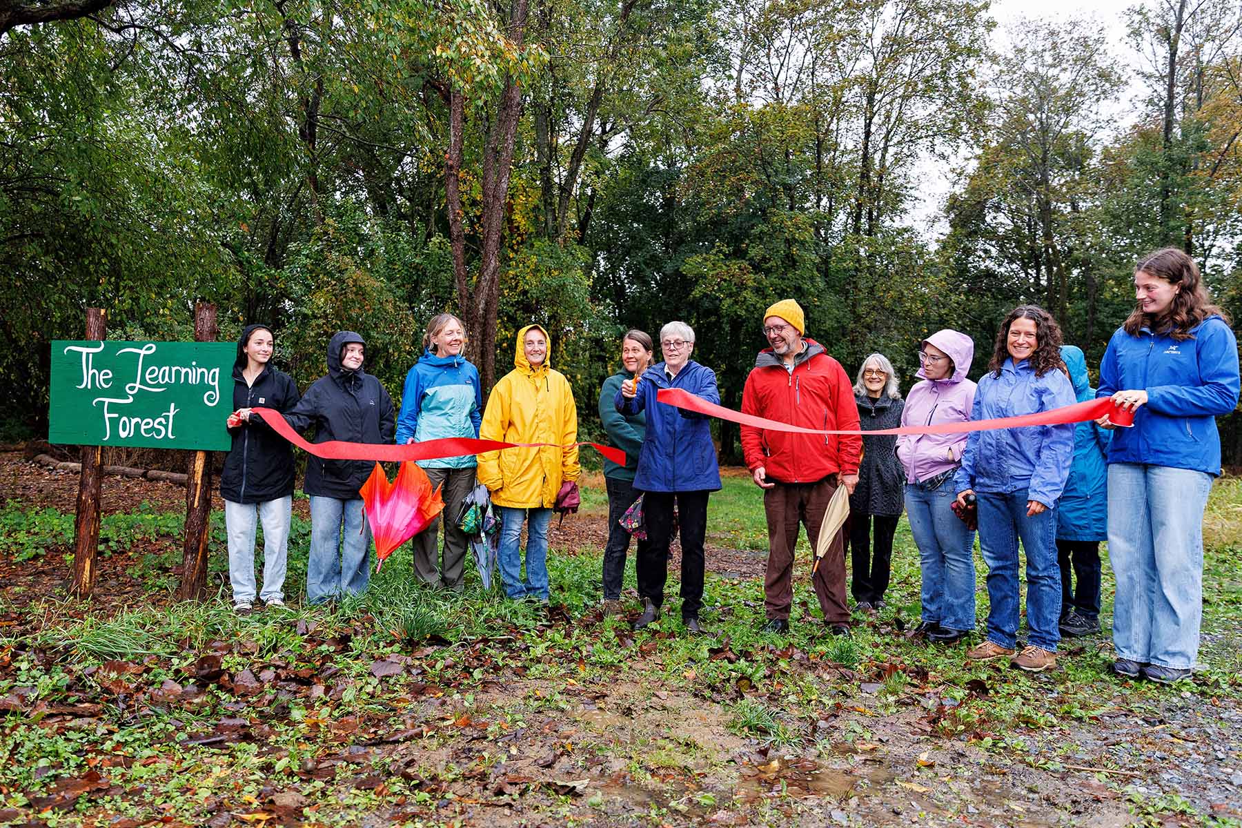 A group of people wearing rain jackets stand outdoors in front of a green sign that reads “The Learning Forest.” They smile as one person cuts a red ribbon during a rainy-day ribbon-cutting ceremony surrounded by trees.