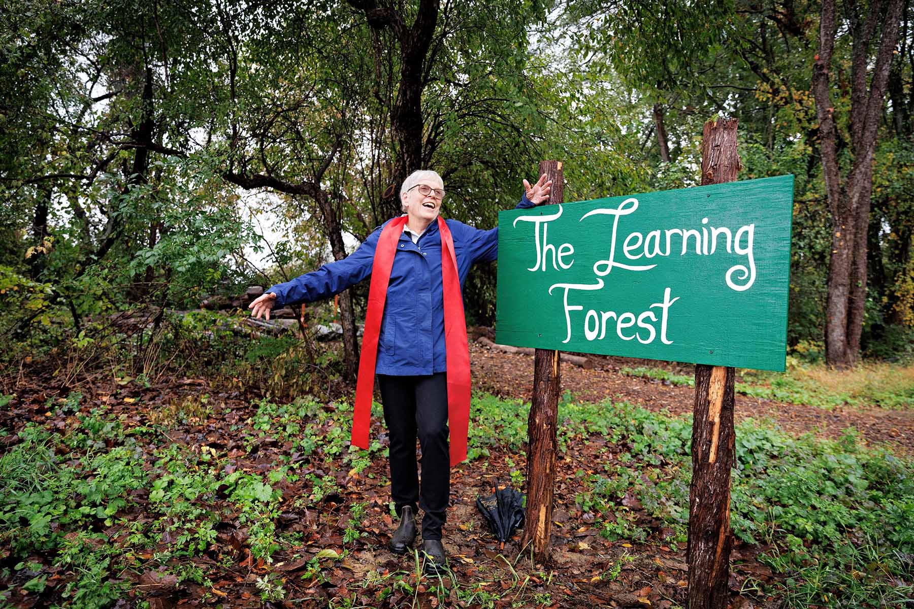 A smiling older woman with short gray hair, wearing a bright blue jacket and a long red scarf, stands with her arms outstretched in a lush, wooded area. To her right is a green wooden sign on two rustic posts that reads "The Learning Forest" in white script.
