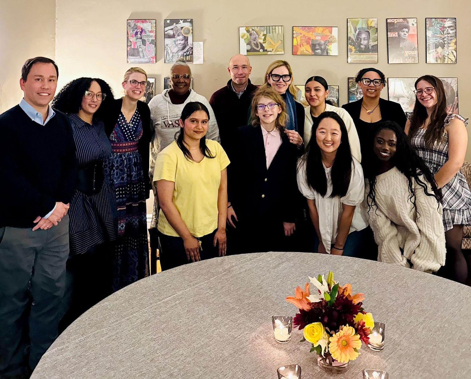 A group of thirteen people stand and smile together in a warmly lit room with artwork on the wall behind them. A round table with candles and a floral centerpiece is in the foreground.