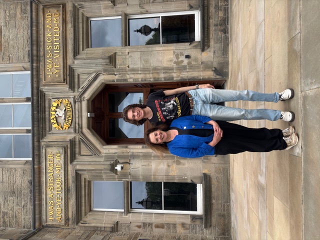 Two people stand in front of a stone building.