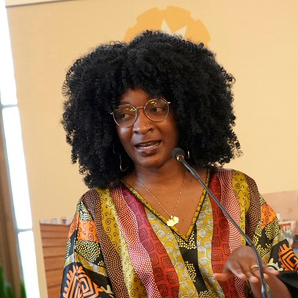 An individual with dark, curly hair and round glasses stands behind a microphone at a lectern, gesturing with one hand. They wear a colorful patchwork-patterned blouse and bracelets. A blurred indoor backdrop shows a banner and window light.