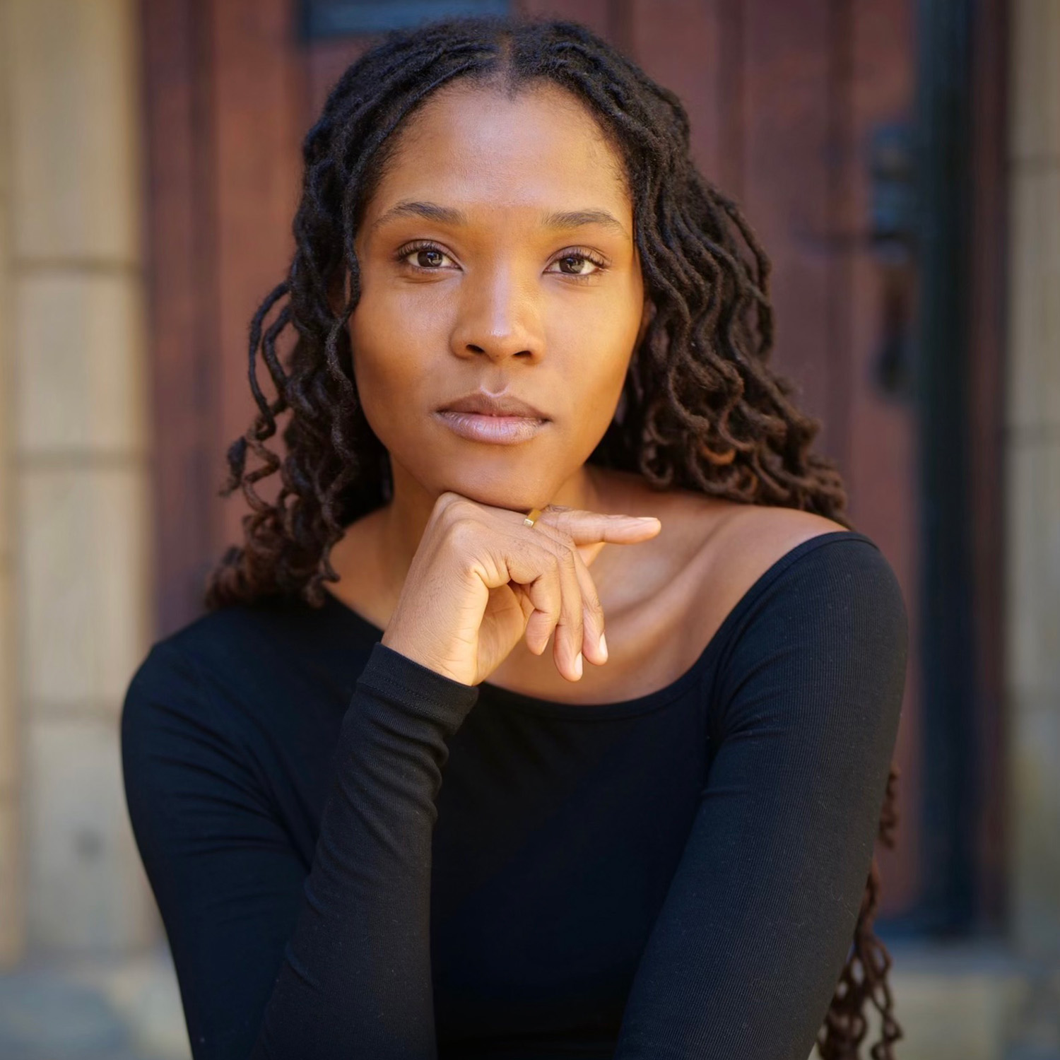Rachel Laryea with long, dark hair wearing a black top with her hand resting under her chin, sitting in front of a wooden door.