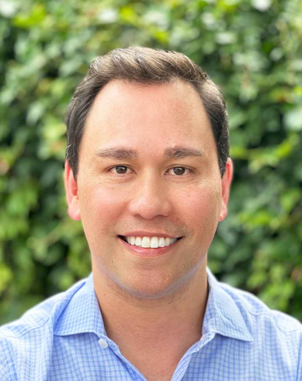 A person with short, dark hair smiles directly at the camera. The person is wearing a light blue checked collared shirt. The background is a softly blurred wall of green foliage.