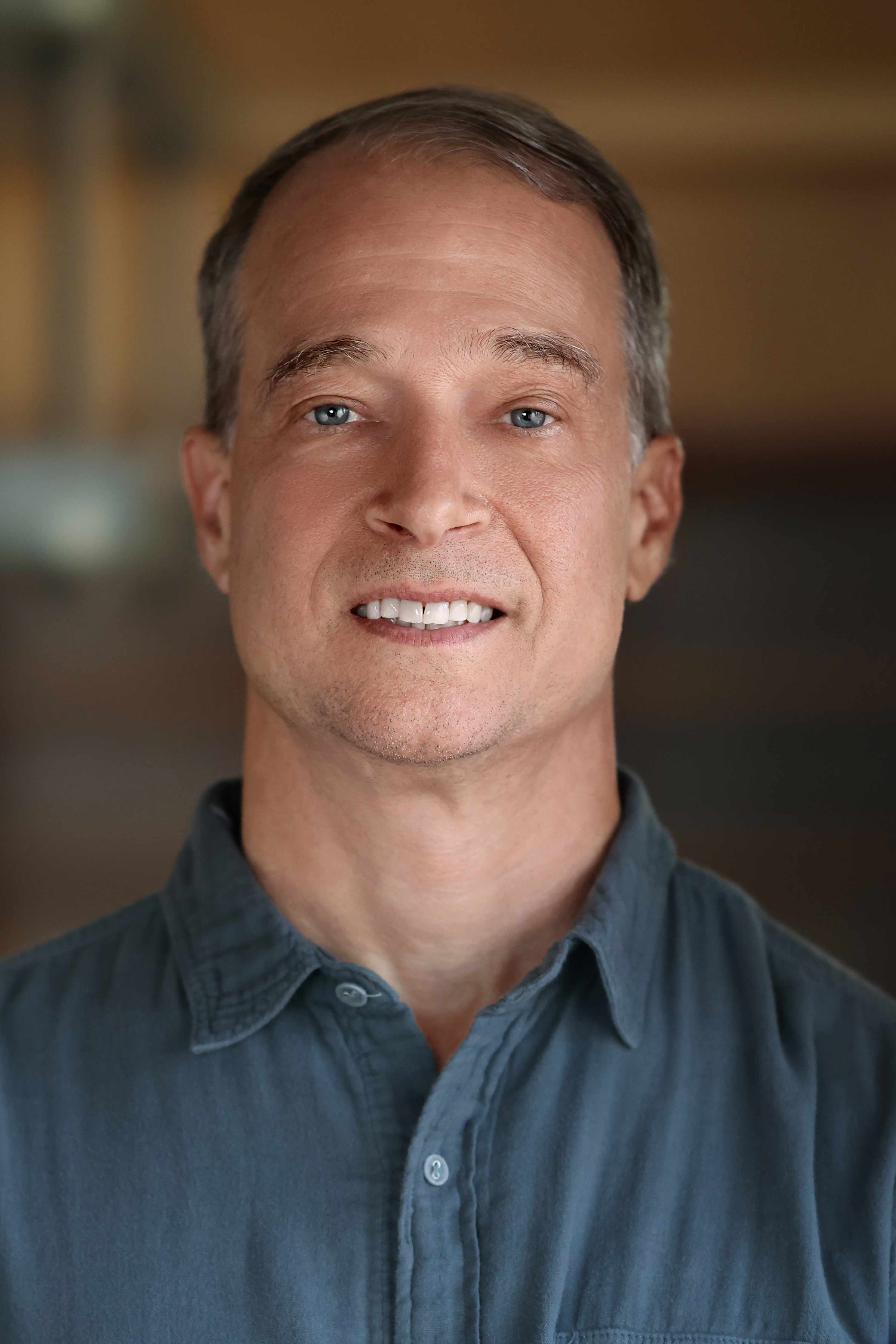  A close-up portrait of a person with short, light brown hair and blue eyes, smiling faintly at the camera. The person is wearing a gray-blue collared shirt. The background is softly blurred with warm, neutral tones.