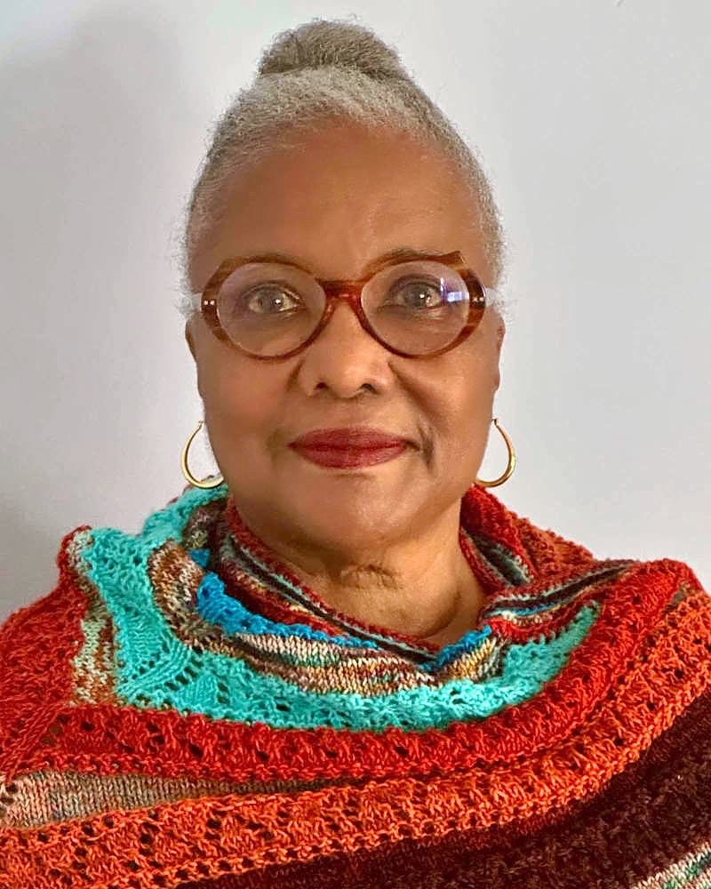 A close-up portrait of a person with light-colored hair pulled back, wearing large oval glasses and gold hoop earrings. The person is looking directly at the camera, wearing a vibrant, multi-colored crocheted or knitted shawl in shades of orange, teal, and brown.