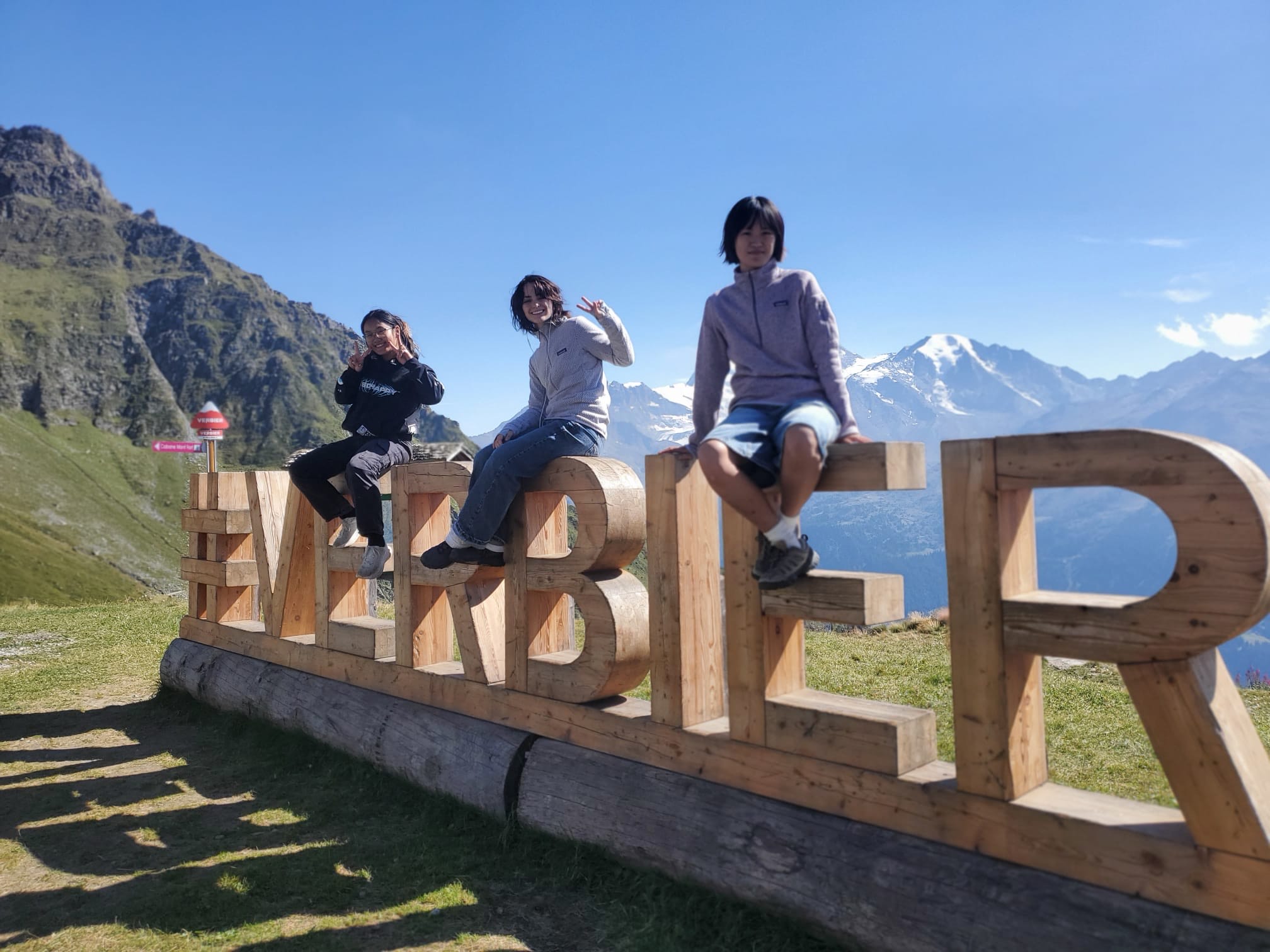 Three students sit outside and smile on top of a sign that spells “#VERBIER.” Mountains are visible in the background.