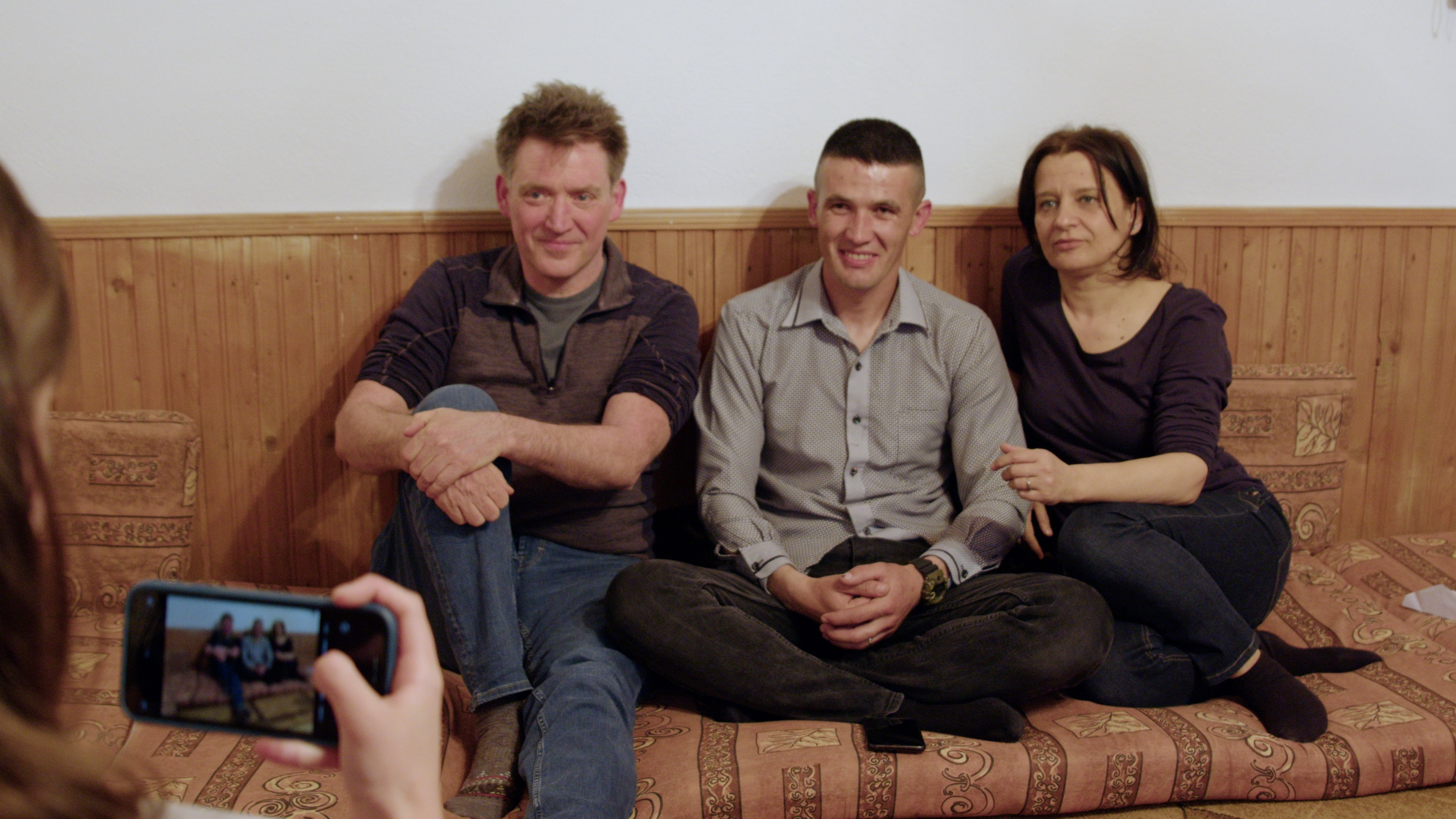 Three people sit on a couch while another person films them with a phone.