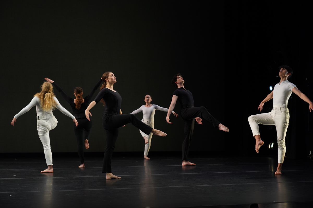 A group of six dancers performs on a dimly lit stage, with a minimalistic black background. The figures are dressed in a mix of black and white clothing, highlighting their contrasting postures and movements. Each dancer exhibits dynamic poses, with arms extended and legs raised, creating a sense of fluidity and grace. The lighting casts subtle shadows on the stage floor, emphasizing the dancers' bare feet and the sleek, polished surface beneath them.