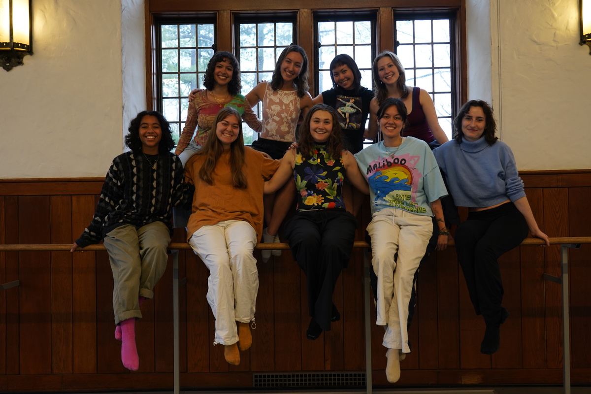 A group of eight figures is arranged on a wooden railing, smiling and interacting in a spacious interior. The scene features large windows in the background that allow natural light to illuminate the warm tones of the wooden walls. Each figure is dressed in a variety of casual clothing styles and colors, creating a vibrant assortment against the muted backdrop. The atmosphere is lively, with a sense of camaraderie evident among the group, highlighted by their varied postures and expressions.