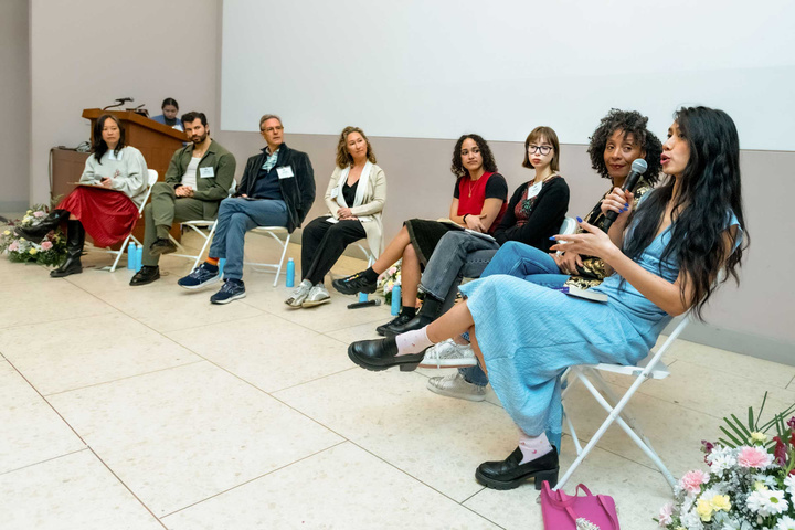 A group of eight individuals sit in a semi-circle on white folding chairs arranged in an indoor setting. In the foreground, a speaker wearing a light blue dress gestures while holding a microphone, her long hair cascading down her shoulders. Each person exhibits a variety of postures and expressions, engaged in discussion; some hold notepads while others listen attentively. The background features a light-colored wall and wooden podium, adorned with floral arrangements at the feet of the participants.