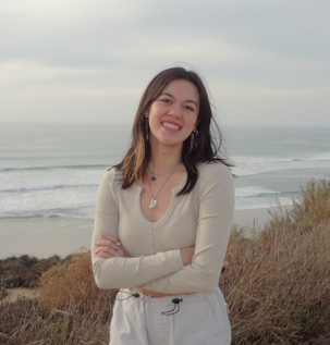 A person stands with arms crossed on a coastal hill, overlooking a beach with gentle waves. The figure, wearing a light-colored long-sleeve top and pants, faces the camera, displaying a relaxed smile. The background features soft, grayish clouds in the sky, creating a calm ambiance, while the muted colors of the ocean blend with the sandy shore and vegetation in the foreground. The scene captures a tranquil coastal landscape, emphasizing the natural beauty of the environment.