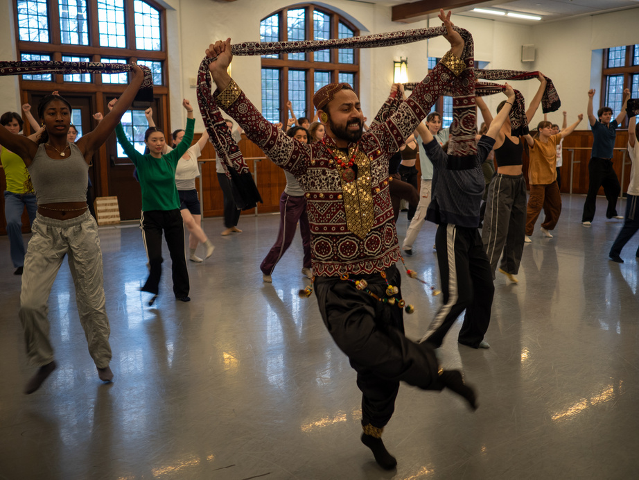 A group of dancers is engaged in a dynamic rehearsal within a spacious studio, characterized by large windows that allow natural light to fill the room. The foreground features a prominent figure in a patterned outfit, adorned with decorative elements, striking a lively pose with arms raised and holding a scarf that flows behind. Surrounding this figure, other dancers are captured in varied postures, some with arms lifted high, creating a sense of movement and rhythm. The walls are a warm wood tone.