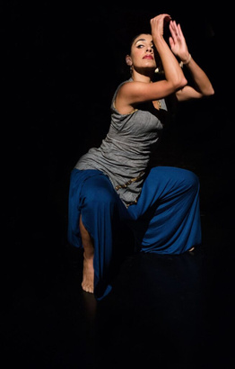A figure poses dynamically in a dimly lit space, emphasizing their elegant lines and form. The outfit consists of a fitted gray top and flowing blue pants, with the fabric creating soft movement around the body. The figure is crouched with one arm raised near the face and the other bent at the elbow, highlighting their expressive posture. A small amount of light illuminates the face, creating sharp contrasts with the surrounding darkness.
