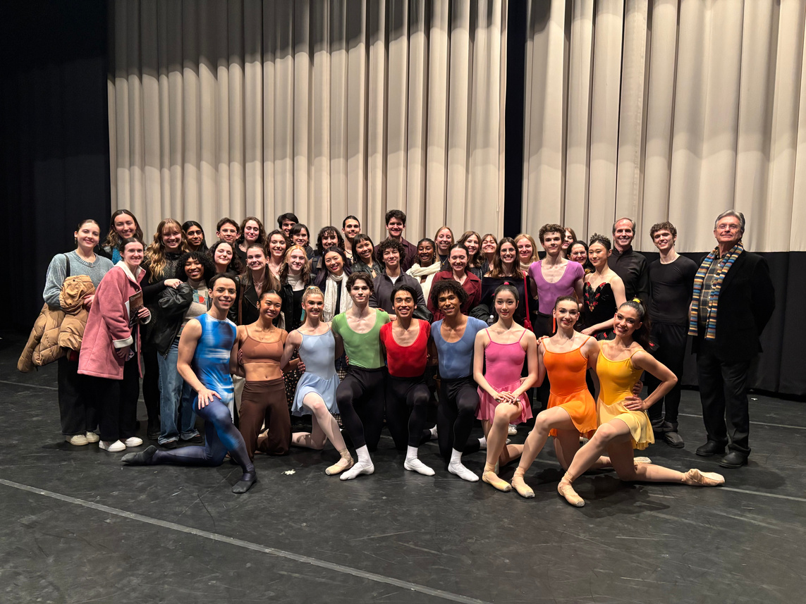 A large group of individuals is arranged in a semi-circle on a stage. The setting features a dark floor illuminated by soft stage lighting, with large fabric drapes in the background. Participants wear a variety of dance attire, including leotards and tights in bright colors, with some individuals in casual clothing. The composition captures a festive atmosphere, showcasing a mix of poses and expressions as they gather together for a memorable photo.
