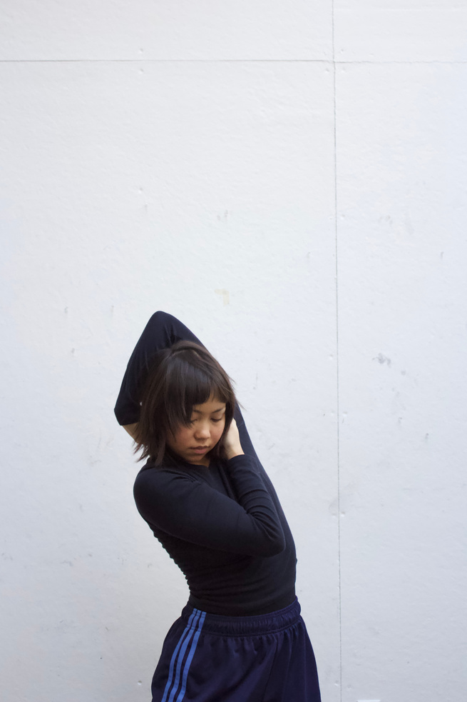 A figure is positioned against a plain, light-colored wall, engaged in a stretching posture with one arm raised overhead and the other arm bent across the body. The clothing consists of a fitted black long-sleeve top and dark pants accented with blue stripes running down the sides. Soft, diffused lighting envelops the scene, creating a calm atmosphere while highlighting the smooth textures of the wall and clothing. The overall composition emphasizes the figure's fluid movement.