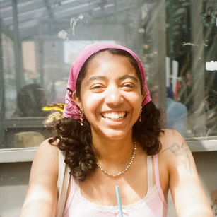 A person sits at a table with a clear glass window behind them, allowing natural light to illuminate their face. They wear a pink headscarf and a light pink top, displaying a bright smile that highlights their features. In their hand, they hold a straw connected to a drink, while their curly hair cascades to the side. The surrounding environment includes blurred figures and hints of greenery, suggesting an outdoor or café setting.