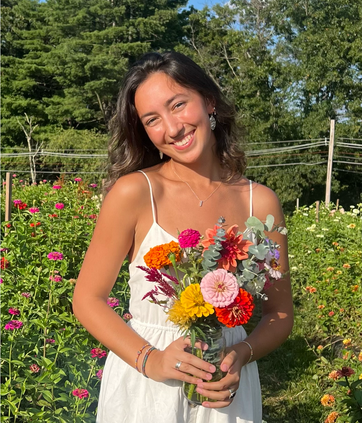 A figure is positioned at the center of a vibrant floral field, surrounded by an array of colorful flowers in various shades of pink, orange, yellow, and red. The figure holds a glass jar filled with a lively assortment of fresh flowers, complementing their attire of a simple white dress with thin straps. Sunlight illuminates the scene, creating a warm glow and casting soft shadows on the ground. Lush green foliage and blurred trees create a natural backdrop, adding depth to the composition.