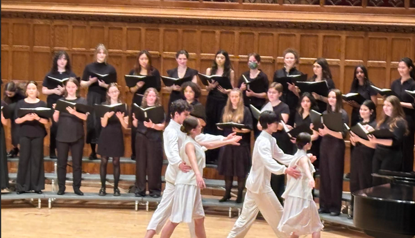 The image features a performance scene set on a tiered stage. In the foreground, two pairs of dancers are engaged in a contemporary dance, with one pair in white clothing and the other in light materials, showing a fluid motion as they connect through their movements. Behind them, a choir is arranged in a semi-circle, dressed in black, holding sheet music and facing the performers, creating a contrast between the active dancers and the static choir. The warm wooden backdrop adds depth to the scene.