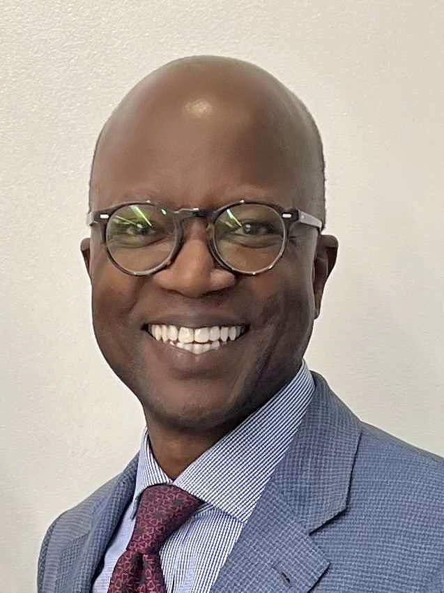  A person in a professional headshot smiles brightly, looking directly at the camera. The person is wearing a light blue patterned jacket, a striped shirt, a dark patterned tie, and round tortoiseshell-frame glasses. The background is a plain, light color.