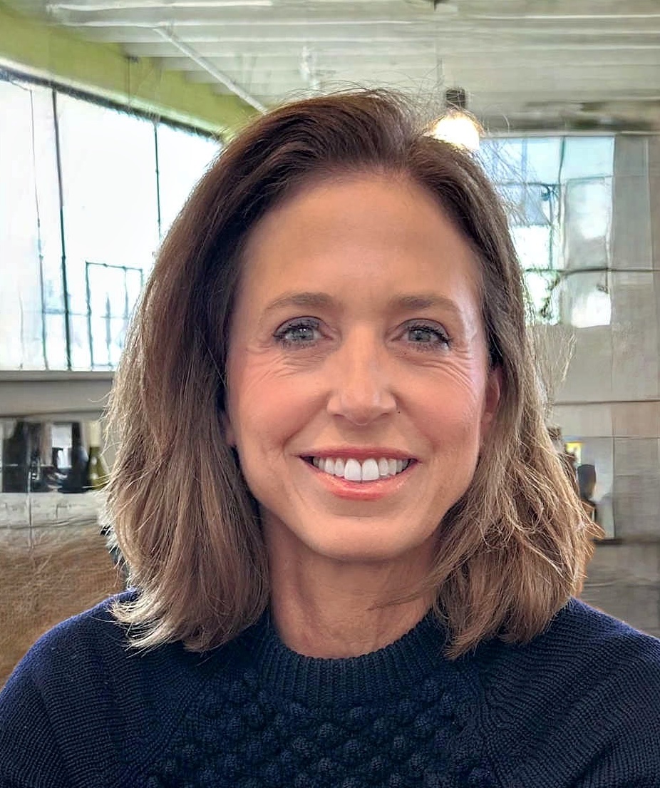 A person with medium-length brown hair smiles directly at the camera. The person is wearing a dark, textured sweater. The background is an indoor setting with large, bright windows and an industrial or loft-style look.