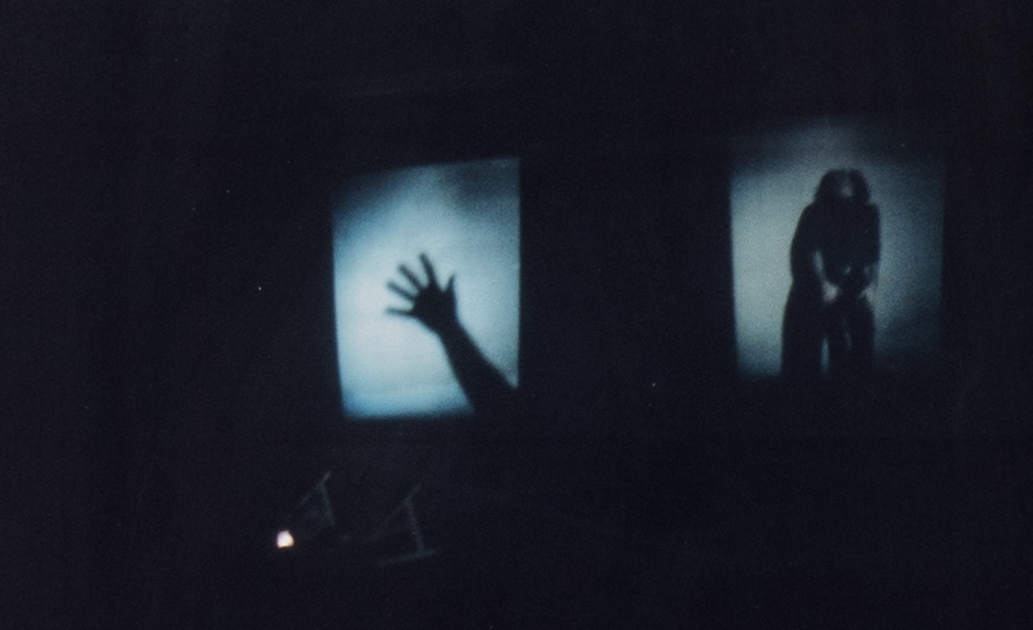 A darkened scene features two illuminated rectangular screens side by side against a black background. The left screen shows the shadow of an outstretched hand and forearm, angled upward with fingers splayed. The right screen displays the shadowy silhouette of a figure bent forward, hands clasped low and head down, partially obscured by darkness. Two small desk lamps sit below the screens, casting faint light upward.