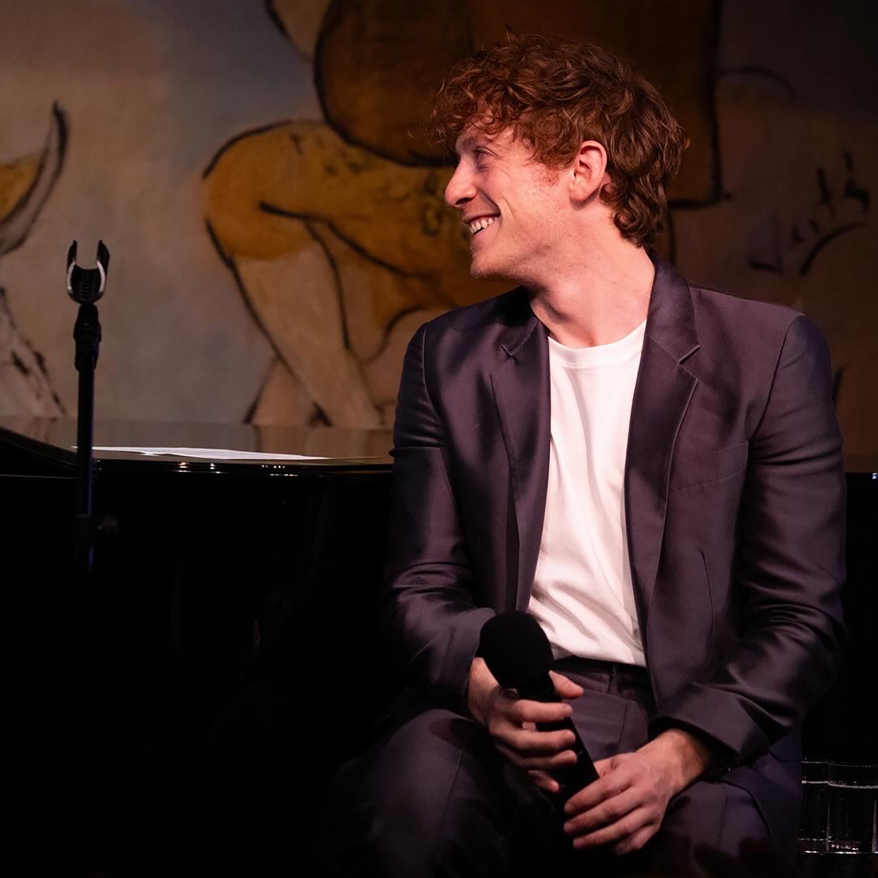 Smiling person seated at a piano holding a mike looking away.