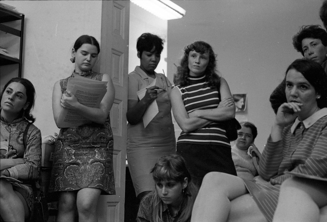 Several individuals gather closely in a small room with plain white walls and a fluorescent ceiling light. Some are seated while others stand, holding or reviewing stacks of typed pages, with one person in the center crossing their arms and another writing on a pad near the doorway. Attentive expressions are visible throughout the group, with a mix of seated and leaning postures. A tall shelf filled with papers stands to the left, and a small framed portrait hangs on the back wall.