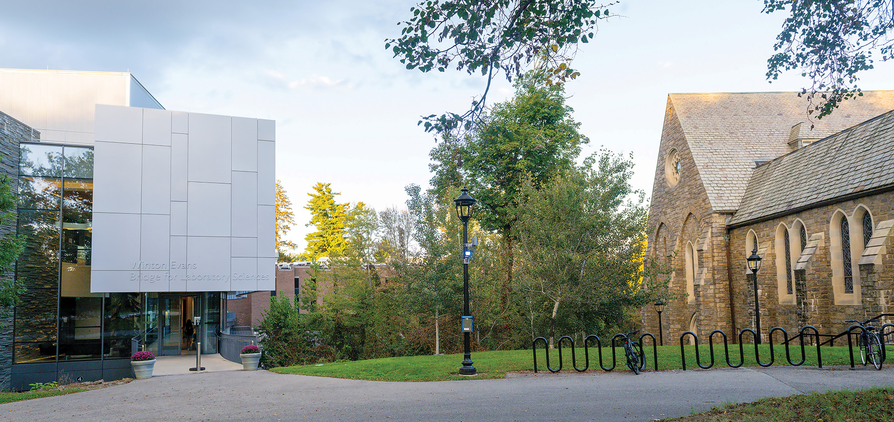 Campus walkway between a modern glass-and-metal building labeled “Winton Evans Bridge for Laboratory Sciences” and a stone chapel, with lampposts, bike racks, trees, and potted flowers in early evening light.