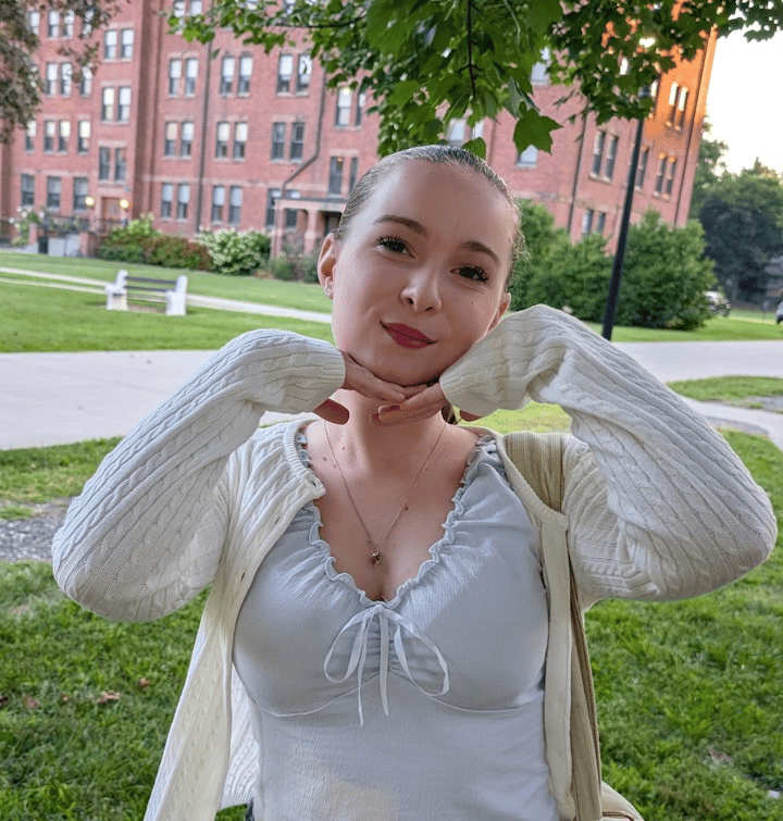 A photo of Alyssa Smith. They are outdoors in front of a brick building, smiling with hands posed under their chin. They wear a light-colored top and cardigan, with trees and grass in the background.
