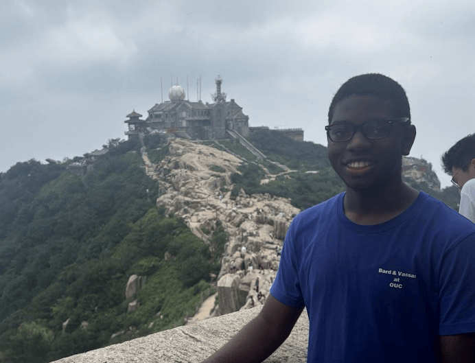 A photo of Aryon Turner, standing on a stone walkway at a mountain viewpoint, smiling toward the camera. A large structure with antennas and domes sits atop the rocky peak in the background under a cloudy sky.