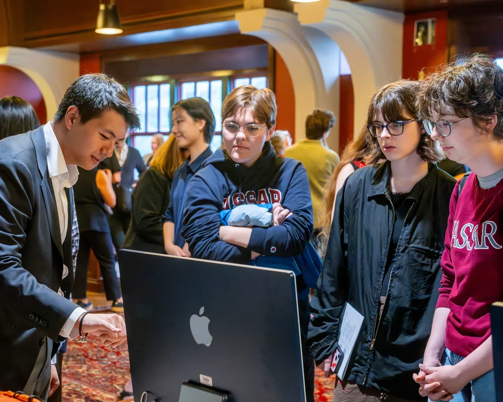 A group of people stand by a person sharing in discussion pointing to a laptop.