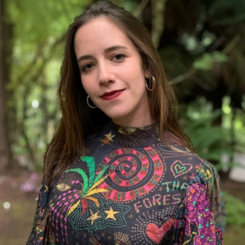  Outdoor portrait of an individual with long dark hair, wearing a high-collared black top with a vibrant, colorful abstract and folk-art-style pattern featuring spirals, stars, and text. They are also wearing silver hoop earrings, against a blurry background of green trees and foliage.